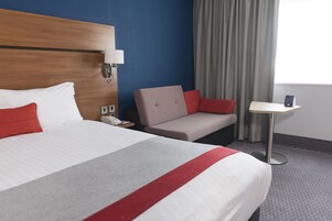 A hotel room with a large bed, a bedside table, a chair, and a small table with a lamp.