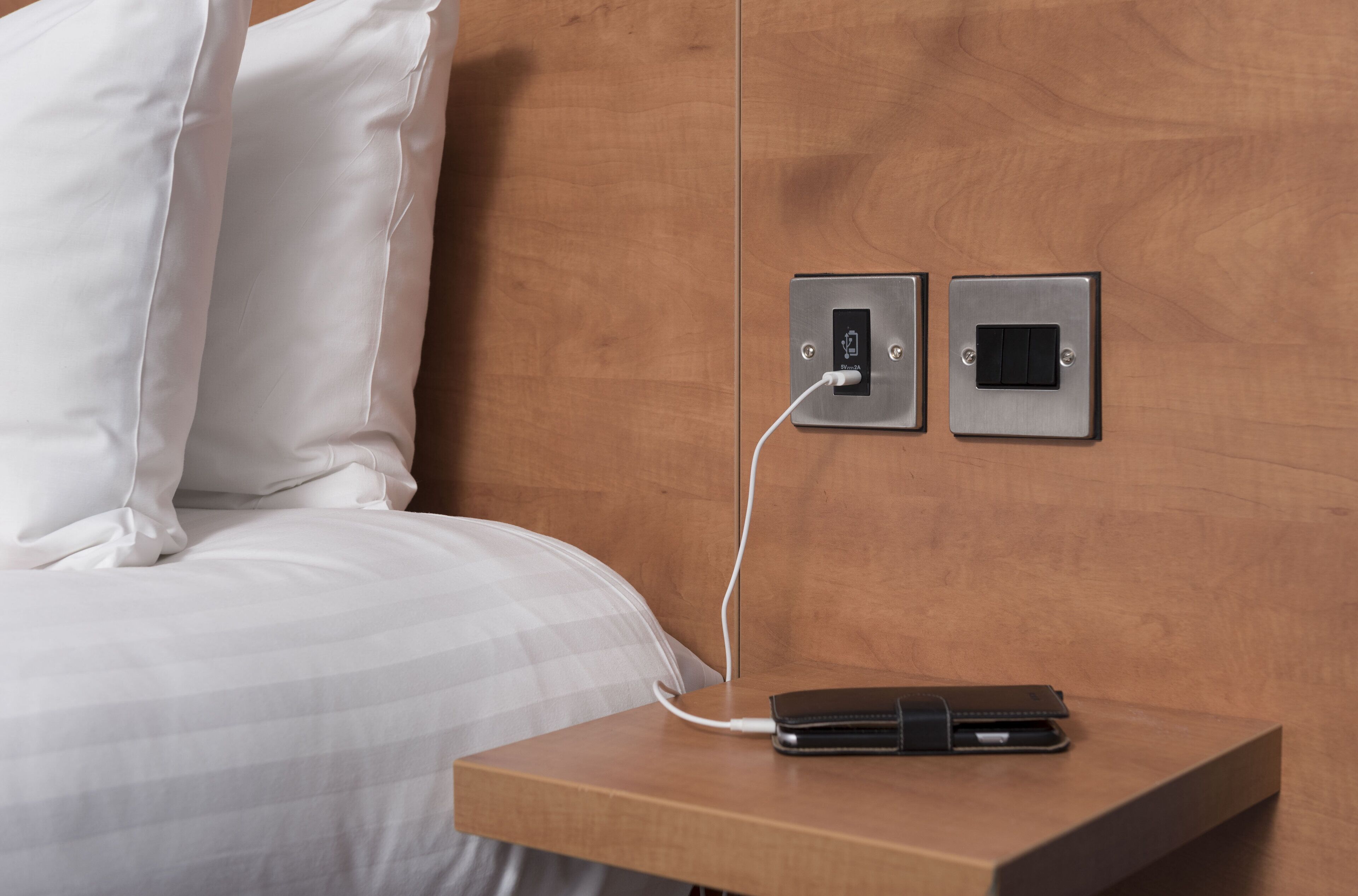 A bed with white linens, a bedside table with a charging cable and a wallet, and two wall-mounted switches.