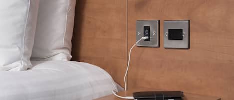 Una cama con sábanas blancas, una mesita de noche con un cable de carga y una billetera, y dos interruptores fijados a la pared.