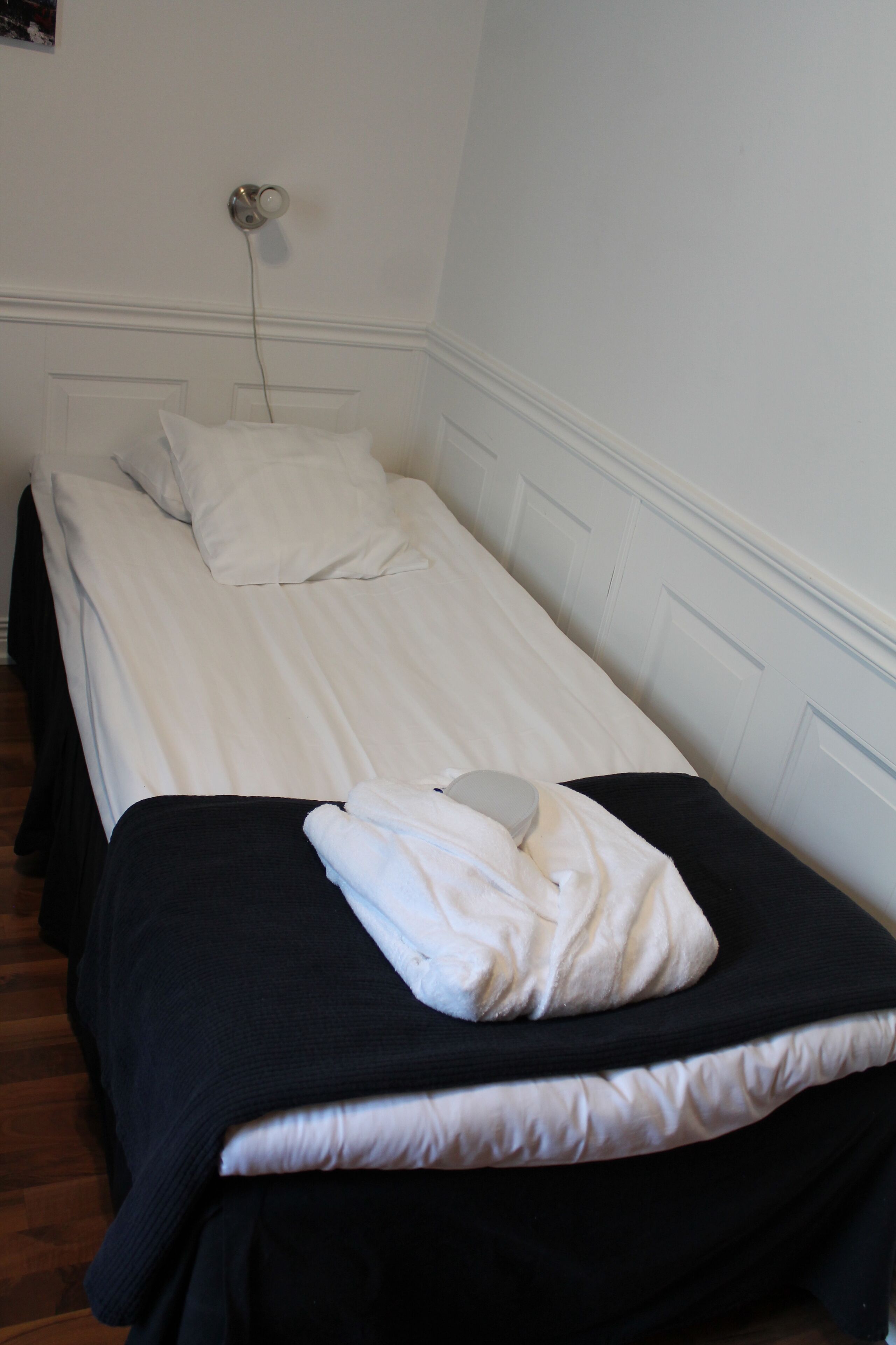 Una cama individual con ropa de cama blanca y un cabecero negro, una almohada blanca y una toalla blanca doblada sobre la cama.