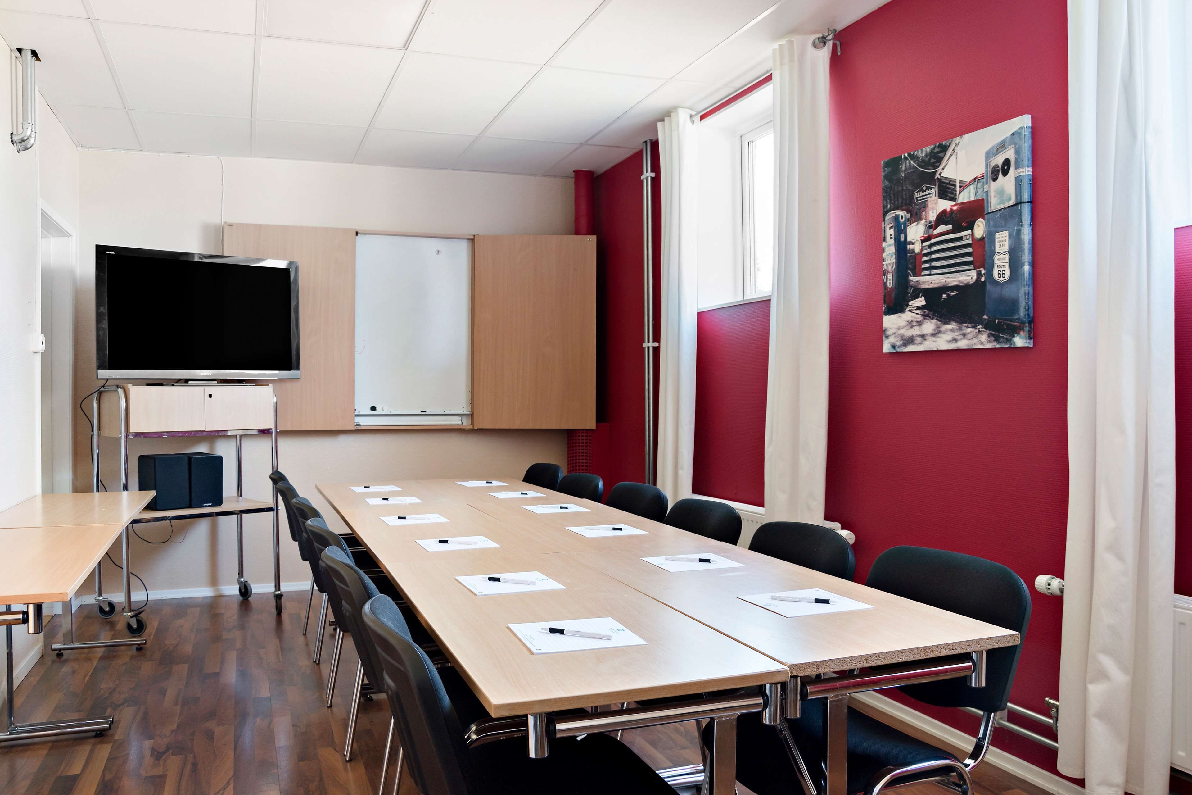 A conference room with a long table, chairs, a TV, a whiteboard, and a red wall with a picture.
