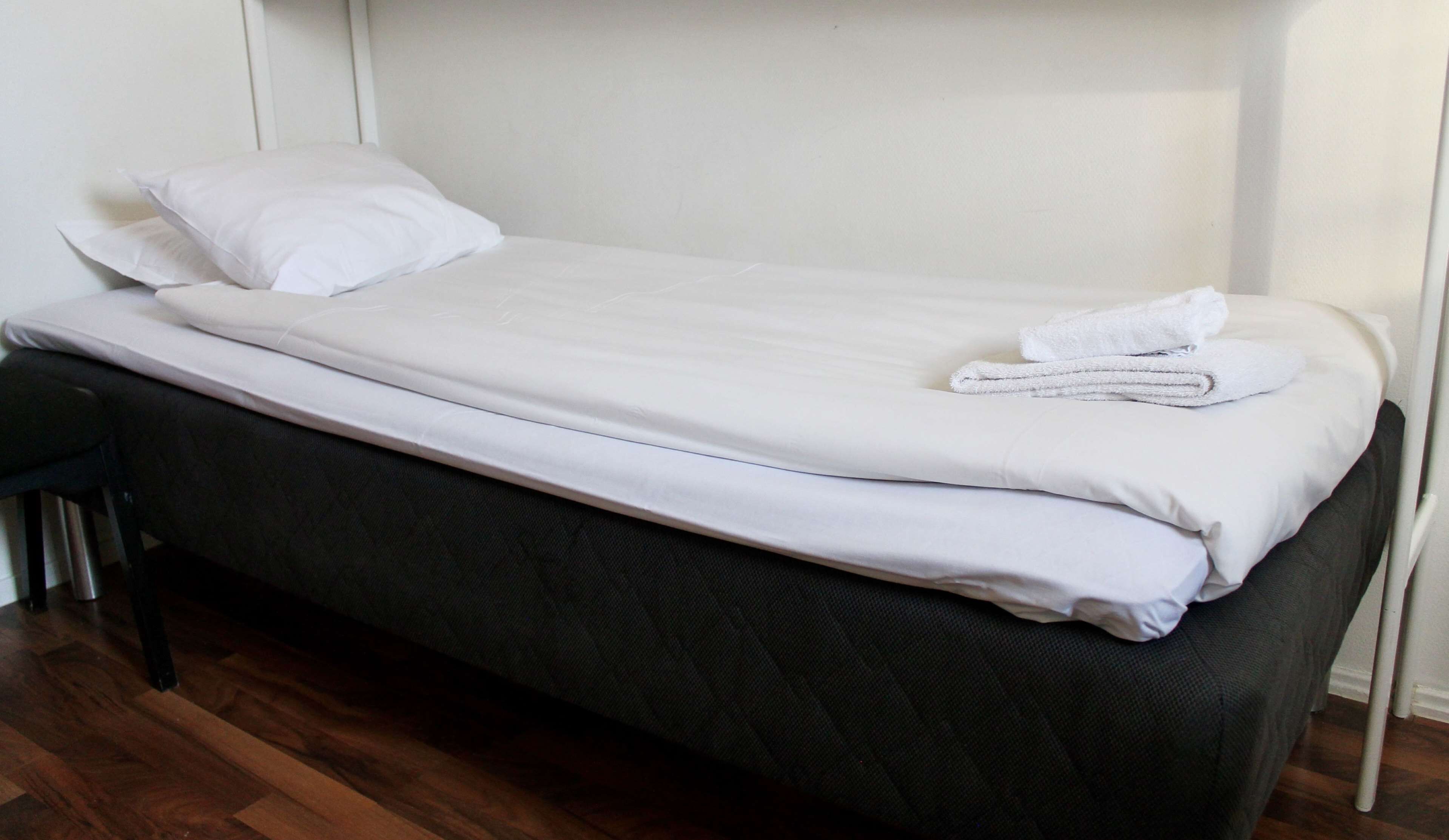 A single bed with white bedding and a folded towel, a black chair, and a wooden floor.