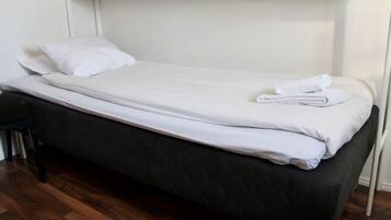 A single bed with white bedding and a folded towel, a black chair, and a wooden floor.