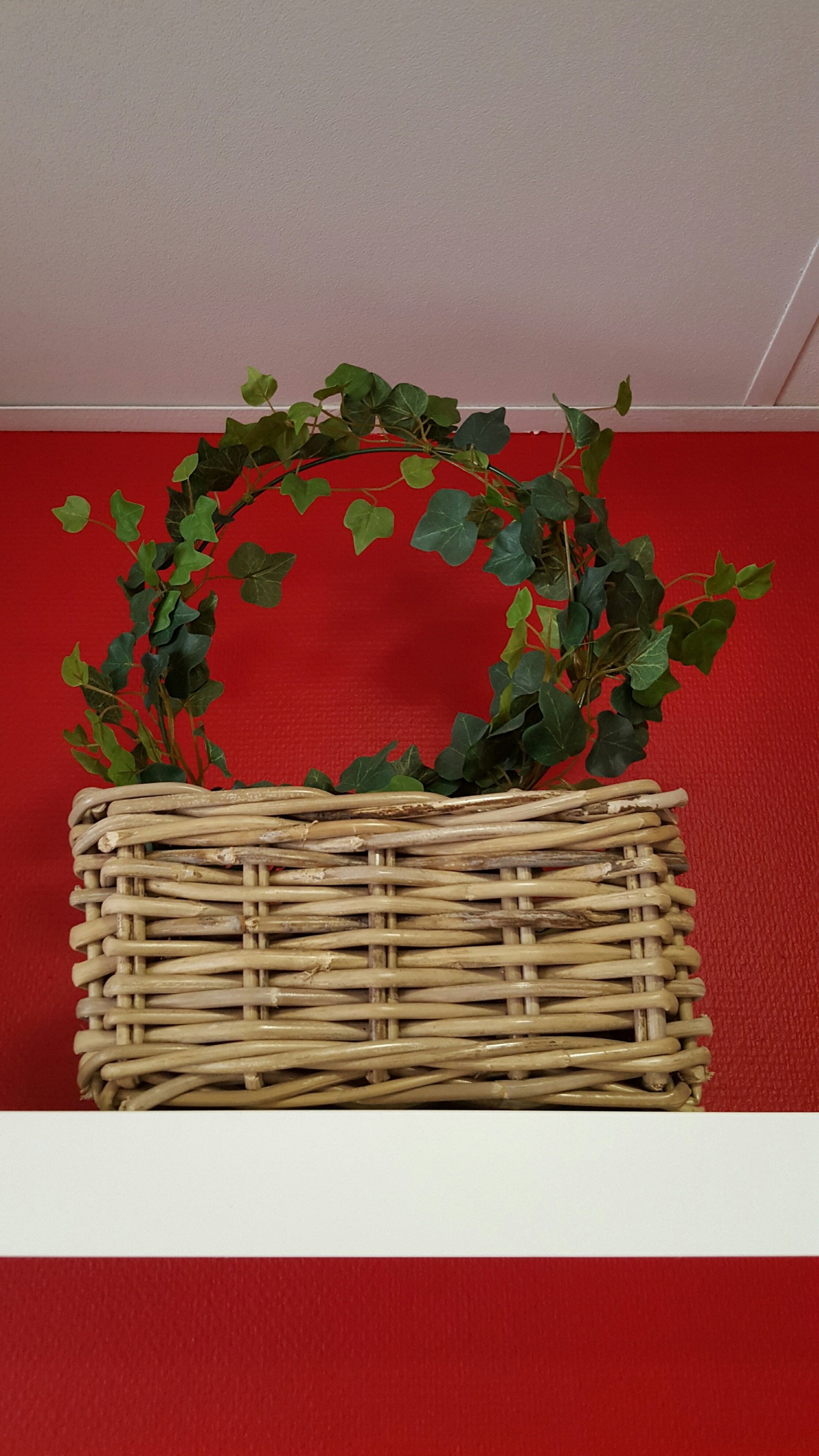 A wicker basket with a green leafy wreath on top, placed against a red background.