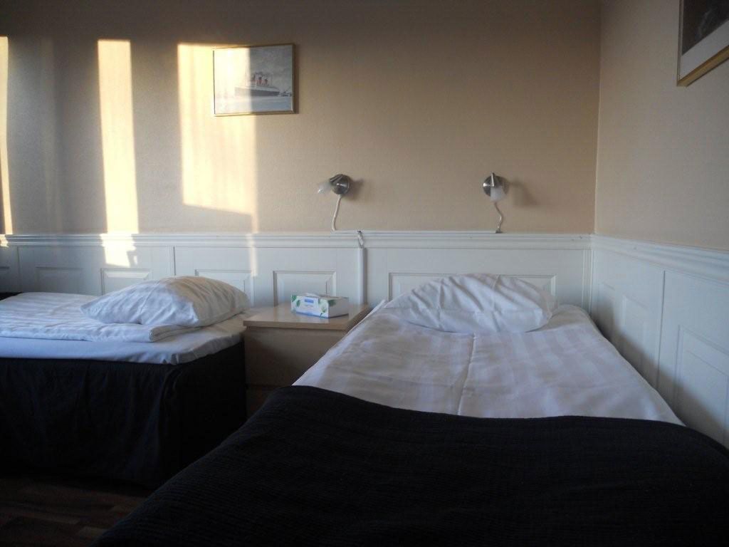 Habitación de hotel con dos camas individuales, una mesita de noche con lámpara, un cuadro en la pared y una ventana con luz solar.