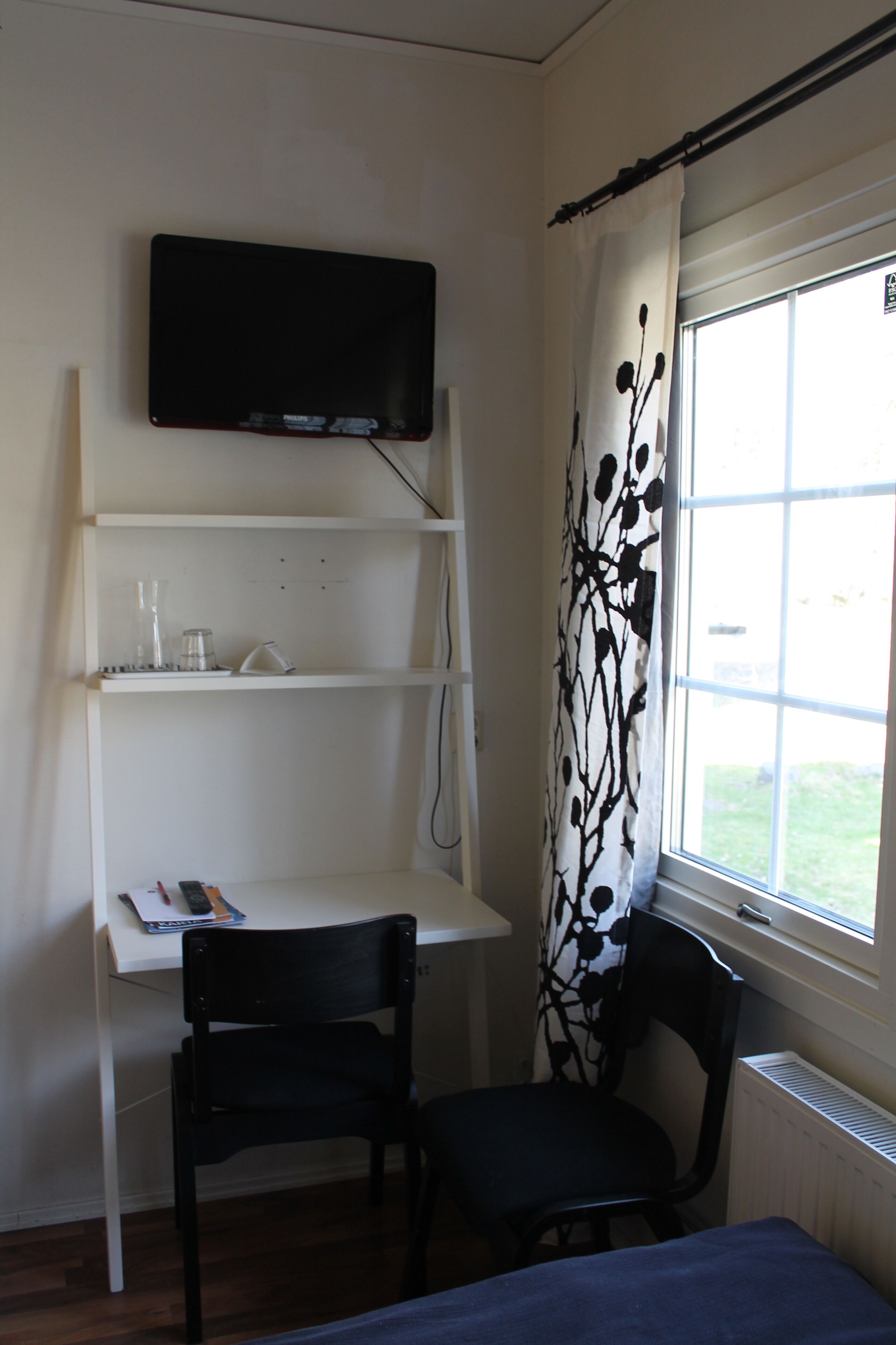 A room with a bed, a desk with a chair, a TV mounted on the wall, and a window with a curtain.