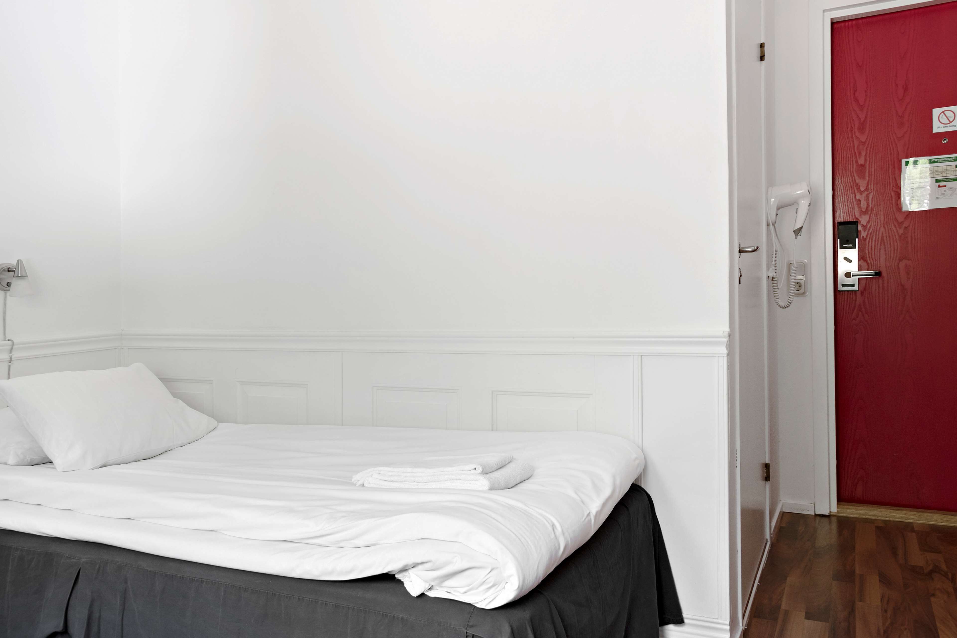A neatly made bed with white linens, a red door with a phone and a sign, and a white wall with a light switch.
