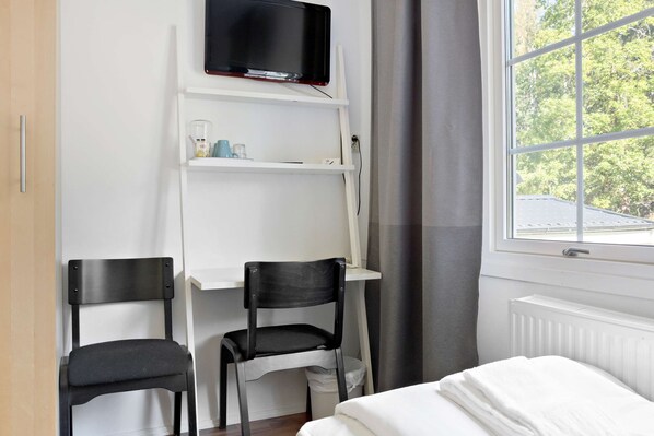 Una habitación con cama, un escritorio con silla, un televisor sobre el escritorio y una ventana con vista a árboles.