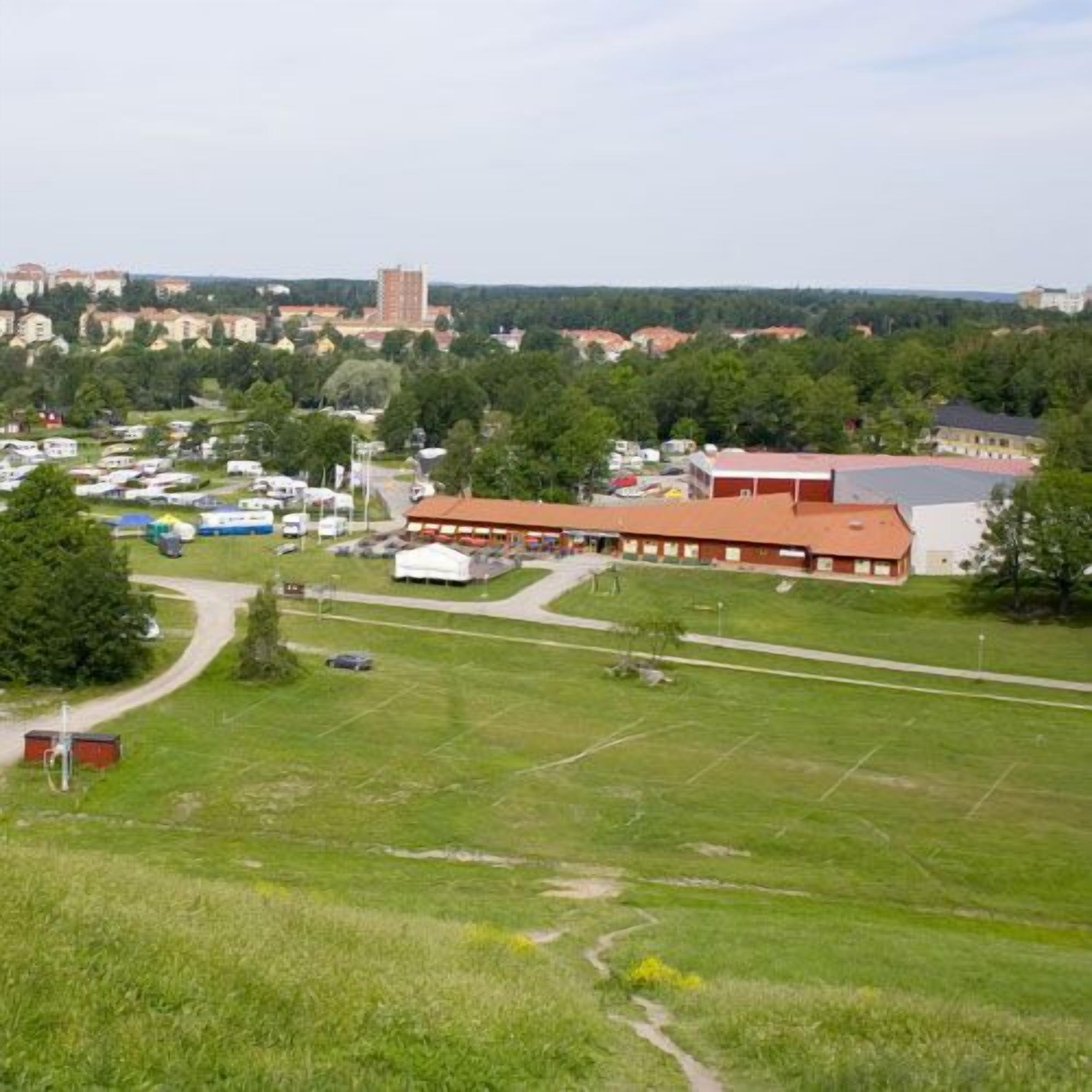 En stor, öppen gräsyta med en byggnad som har rött tak, omgiven av träd och andra byggnader i bakgrunden.