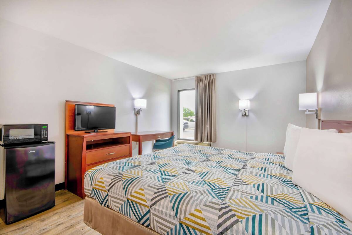 A hotel room with a bed, a desk, a television, a refrigerator, and two wall-mounted lamps.