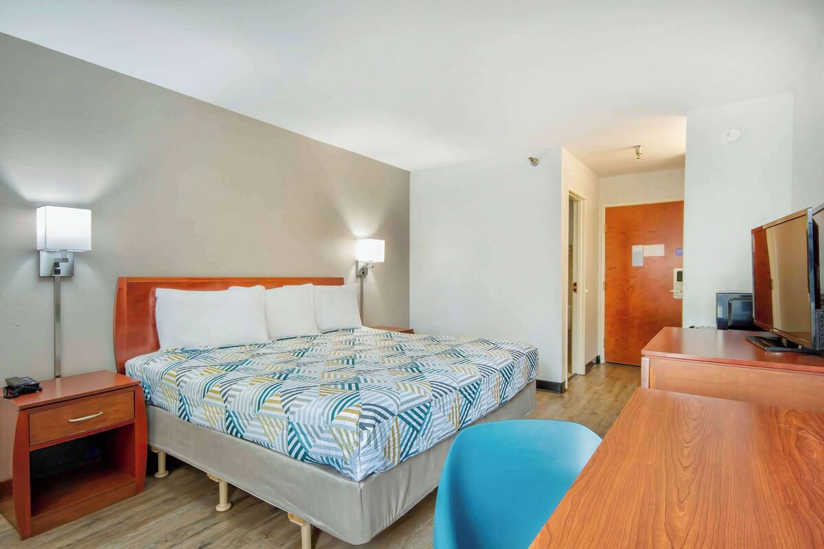A hotel room with a bed, bedside table, lamp, desk, chair, and a television.