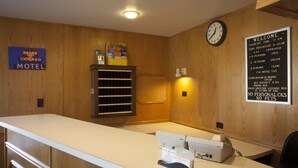 Un comptoir d’accueil d’un motel, avec une horloge, une pancarte de bienvenue et un panneau indiquant les différents types de chambres et leurs tarifs.