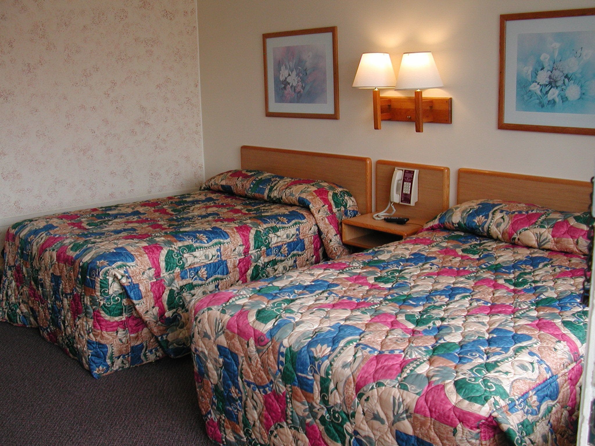 A hotel room with two beds, floral-patterned bedding, a nightstand with a phone, and two wall-mounted lamps.