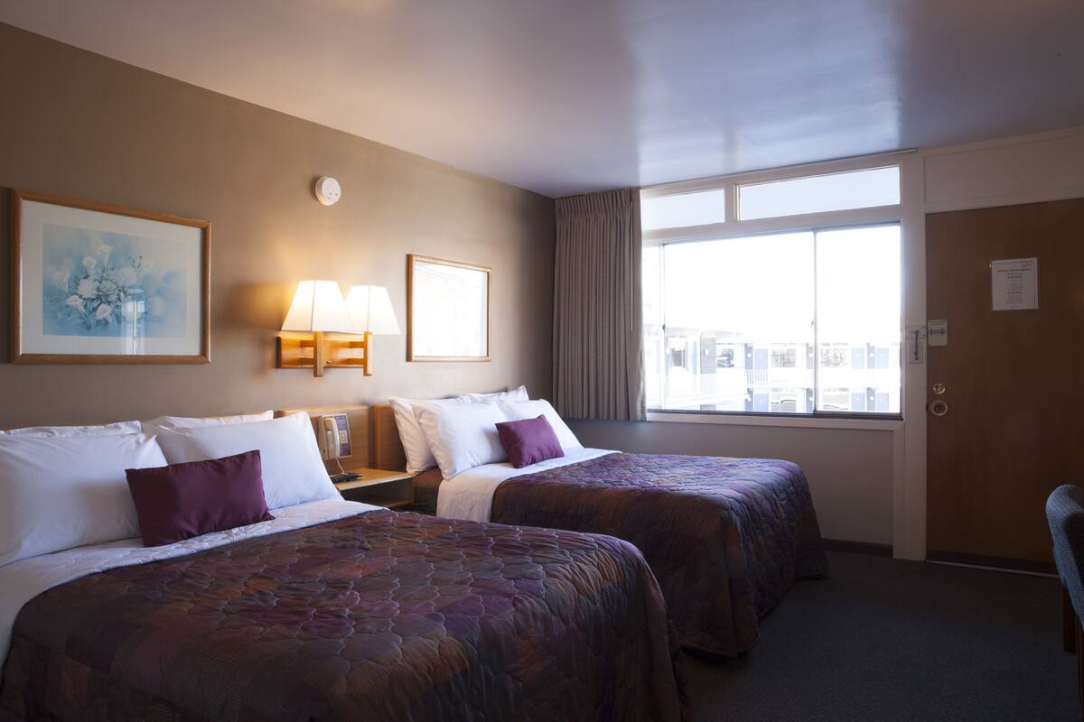 Habitación de hotel con dos camas, un ventanal grande con vista a la ciudad, un cuadro en la pared y una lámpara en la mesita de noche.