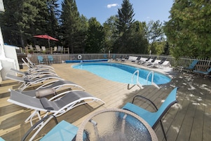 Zona de piscina con sillas de descanso y una mesa, rodeada por una terraza de madera y una valla metálica.