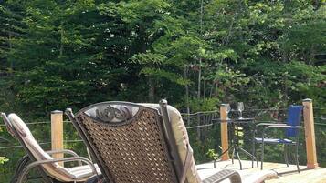 A wooden deck with wicker chairs and a table, surrounded by dense green foliage.