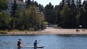A calm lake with two people rowing a canoe, surrounded by trees and a building in the background. - Auberge Hôtel Spa Watel (Sainte-Agathe-des-Monts)