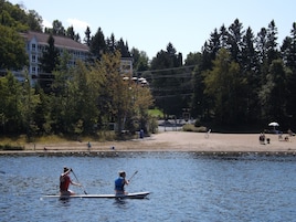 Un lago tranquilo con dos personas remando en un canoa, rodeado de árboles y un edificio al fondo.