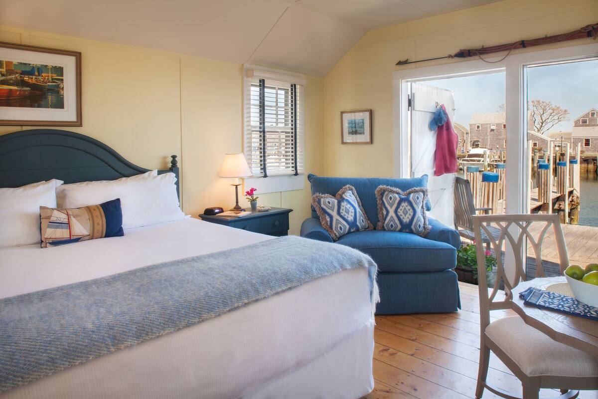 A hotel room with a bed, a blue armchair, a wooden chair, and a view of the outside.