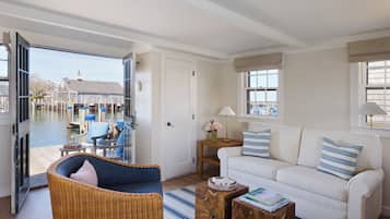 Sala de estar com sofá branco, almofadas listradas em azul e branco, uma poltrona de vime e uma mesa lateral de madeira com livros.