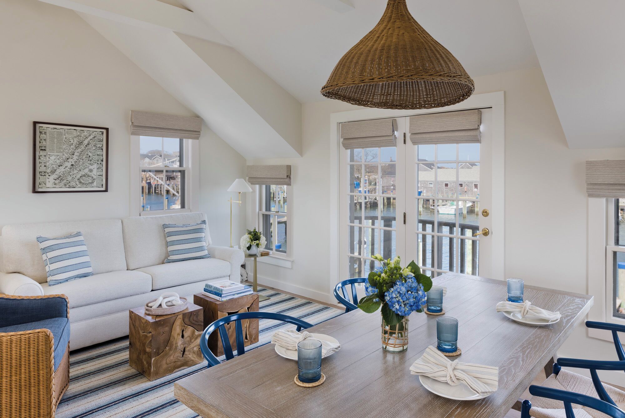 A spacious living room with a high ceiling, a white sofa, a dining table set for four, and a view of a water body through large windows.