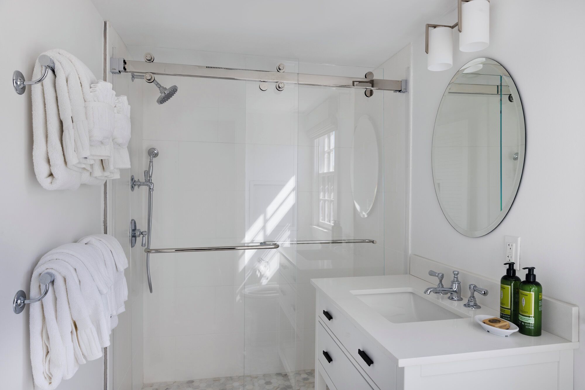Un baño moderno con cabina de ducha de vidrio, tocador blanco con lavabo y espejo, y toallas ordenadas en un perchero.