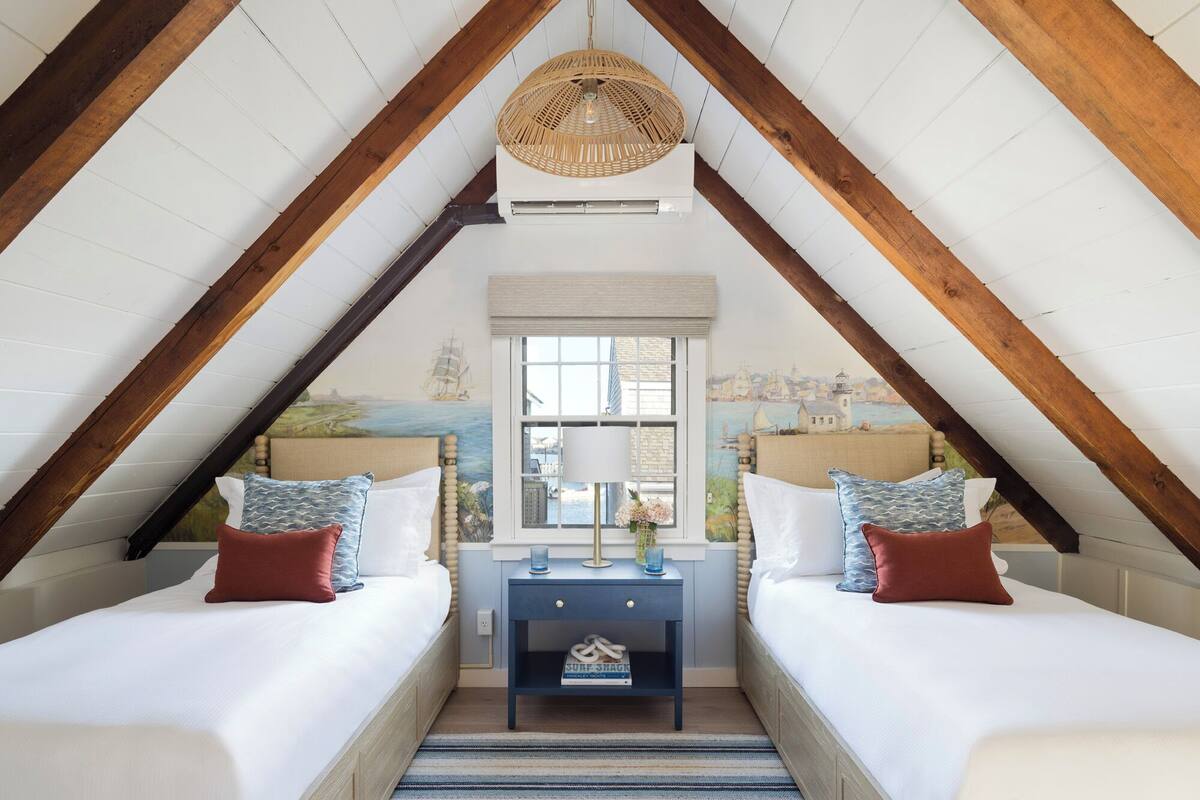 A cozy attic bedroom with two beds, a window with a view of a coastal scene, and a hanging light fixture.