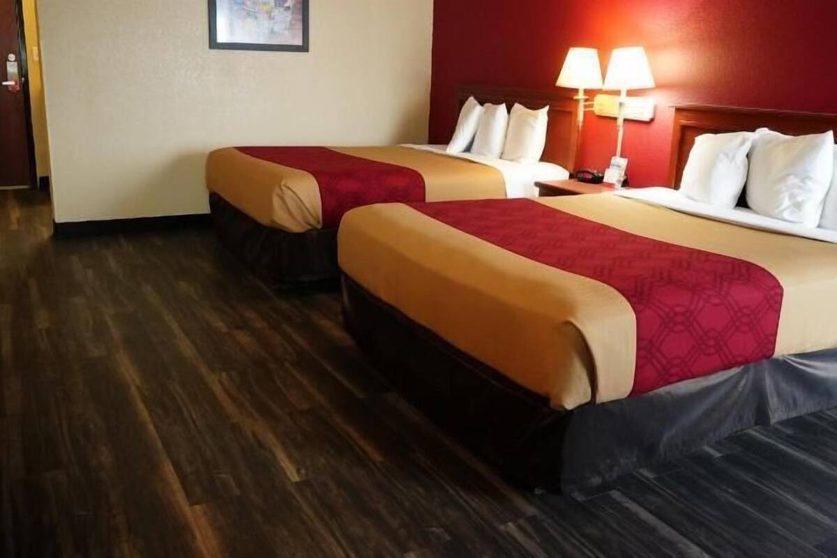 A hotel room with two beds, a wooden floor, a red wall, and a nightstand with a lamp.