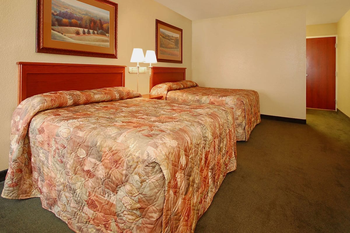 A hotel room with two beds, a nightstand with lamps, and framed landscape paintings on the wall.
