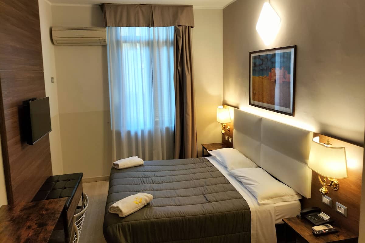 A hotel room with a bed, a bedside table, a lamp, a painting, and a television on the wall.