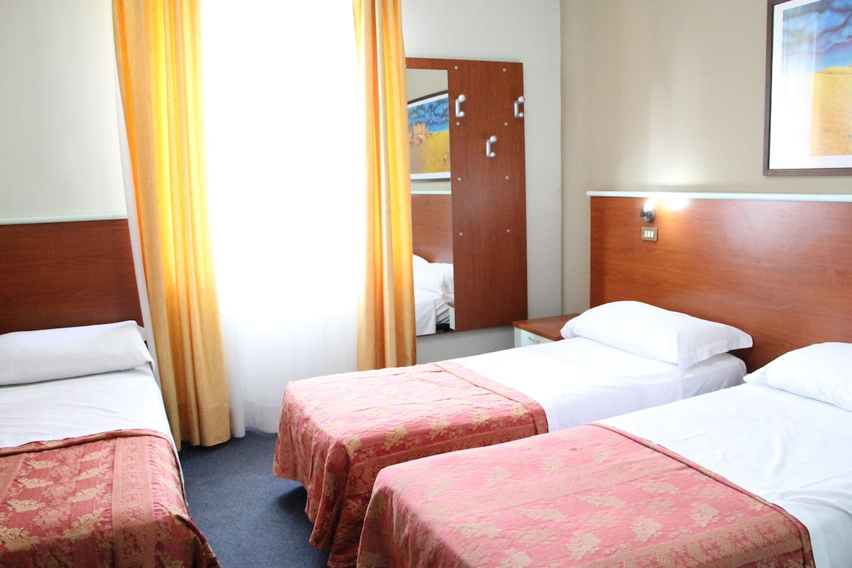 A hotel room with three single beds, each with a wooden headboard and a floral bedspread. There is a wooden wardrobe with a mirror and a framed picture on the wall.