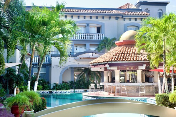 Un área de piscina con bar, sillas de descanso y un edificio con tejado de azulejos y balcones.
