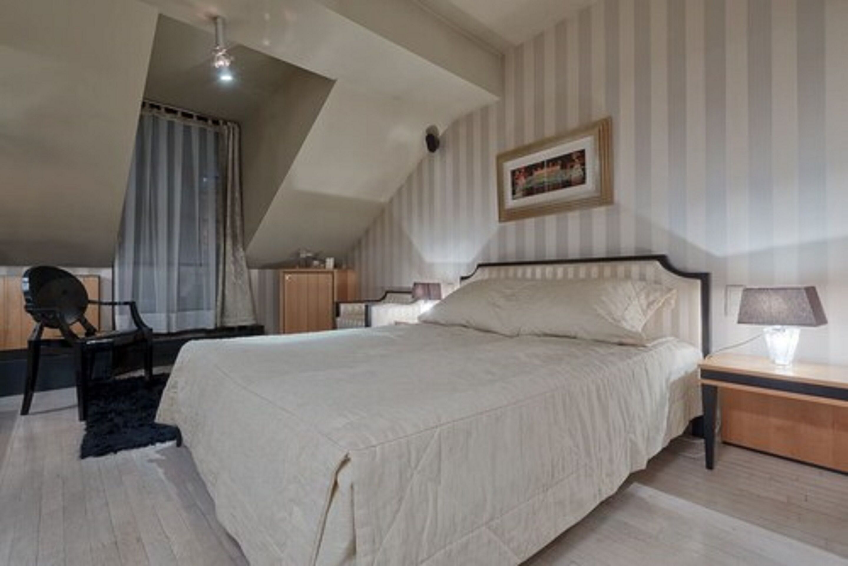 Un dormitorio con cama, una silla, una mesita de noche, una lámpara y un cuadro en la pared.
