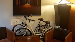 A hotel room with a large painting of an archway, two bicycles, a table with a glass, and a lamp.
