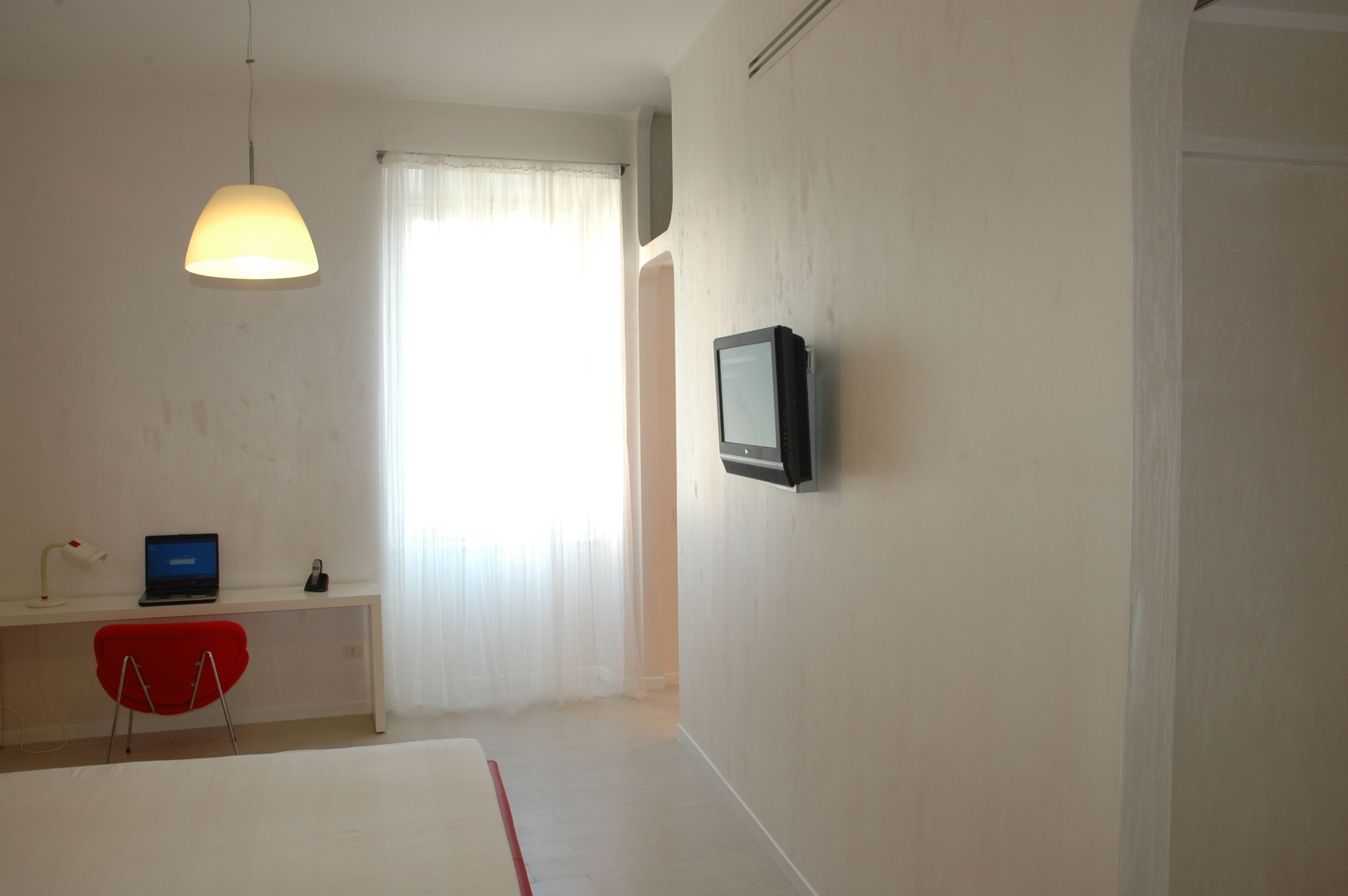 Ein Schlafzimmer mit einem Bett, einem Schreibtisch mit Laptop, einem roten Sessel, einem an der Wand befestigten Fernseher und einem Fenster mit transparenten Vorhängen.