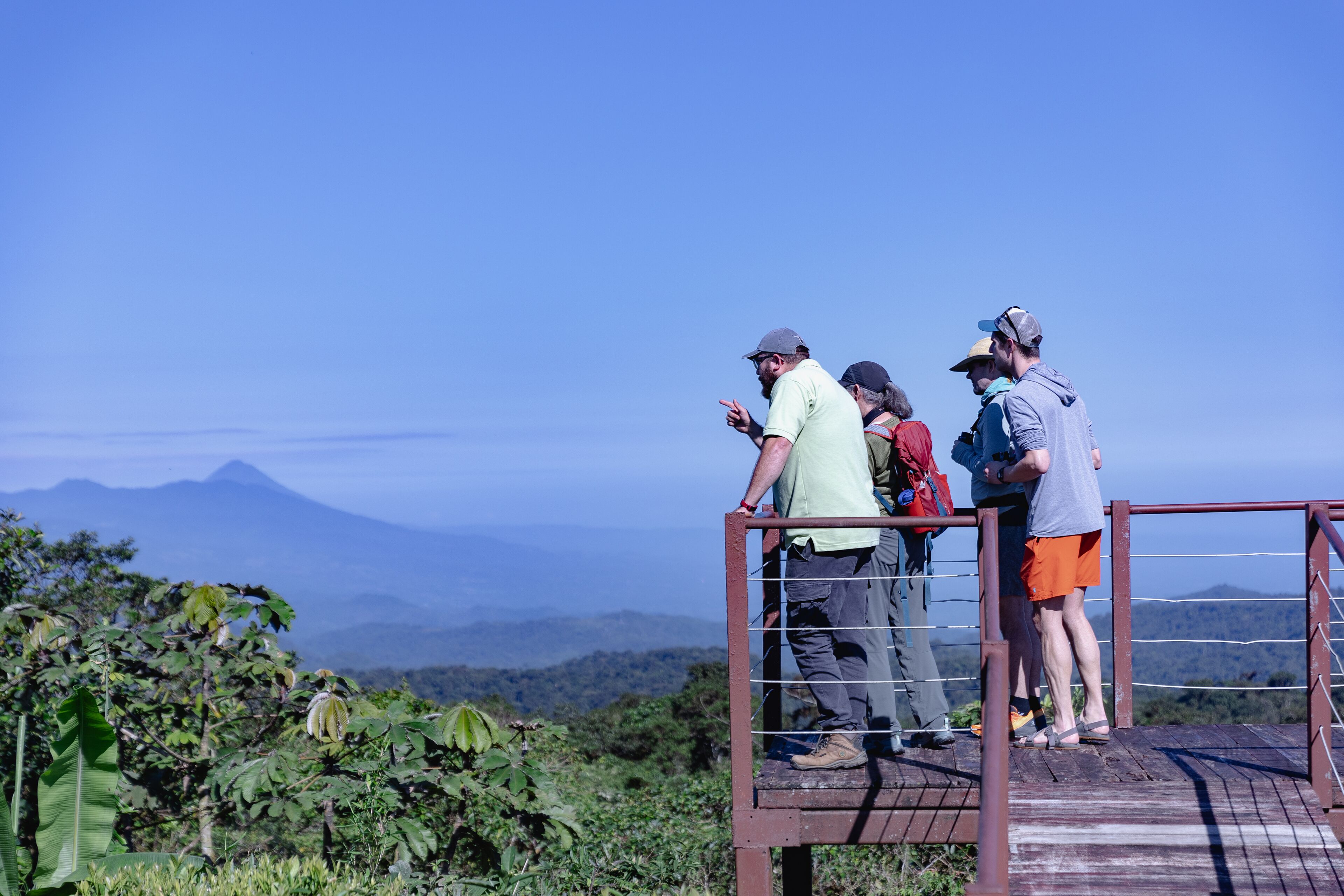 A group of people standing on a wooden platform with a metal railing, overlooking a mountainous landscape.
