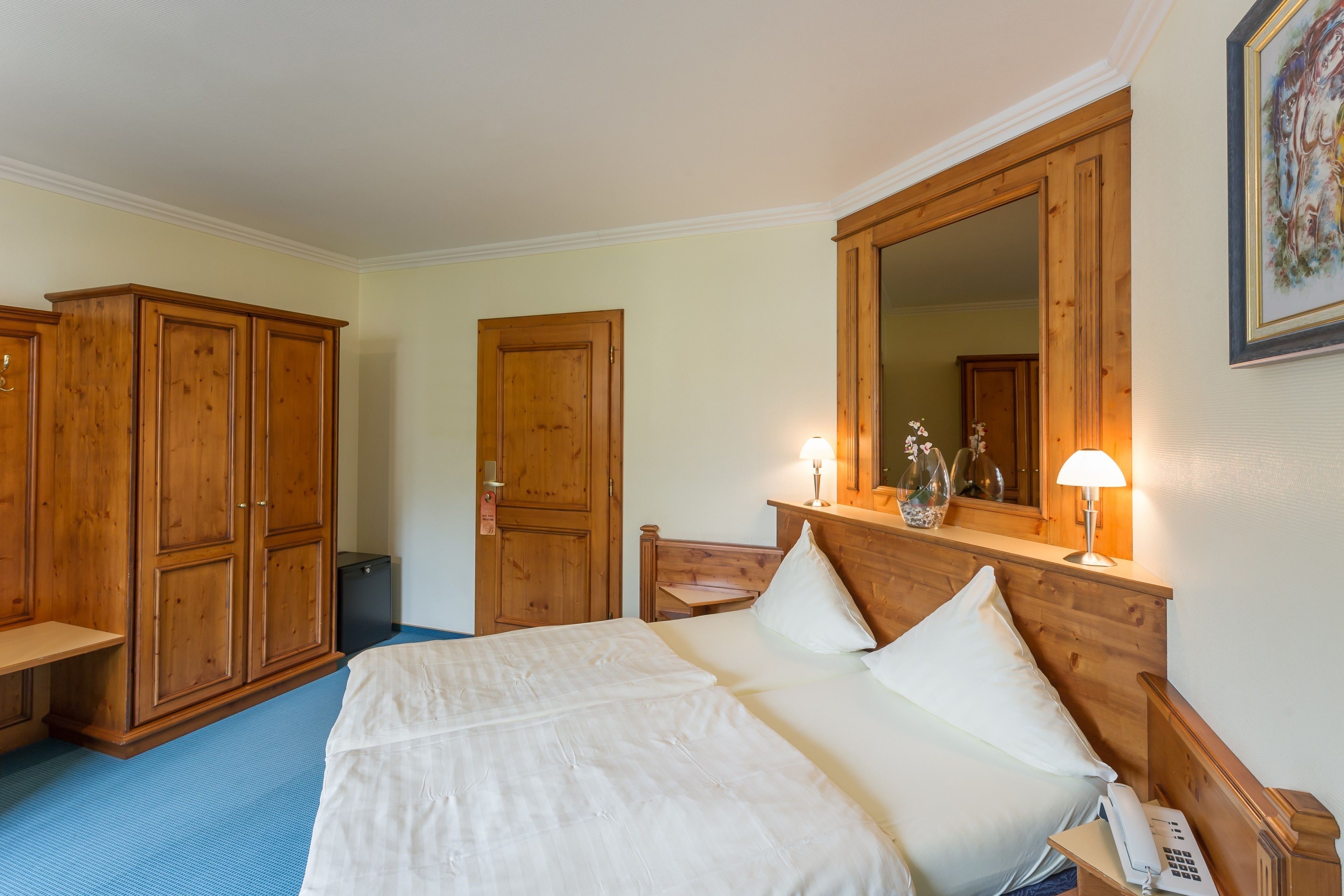 A bedroom with a large bed, wooden wardrobe, a desk with a lamp, and a mirror above the bed.