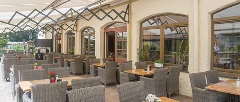 An outdoor cafe with wicker furniture, wooden tables, and potted plants.
