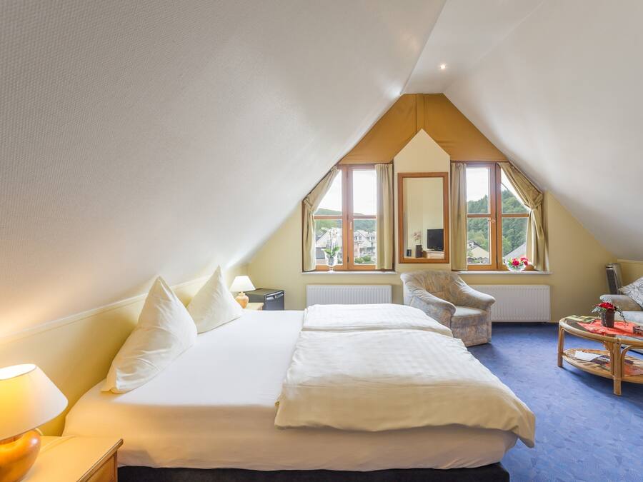 A bedroom with a large bed, a sitting area with a chair and table, and a window with a view of greenery.