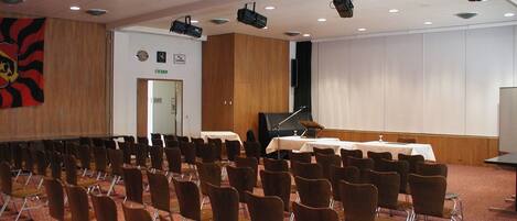 Une salle de conférence avec des rangées de chaises, une scène avec un piano et un grand écran.