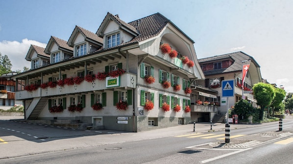 Un hotel de estilo arquitectónico europeo tradicional, con una entrada destacada que incluye una escalera, múltiples ventanas con contraventanas verdes y jardineras en la fachada.