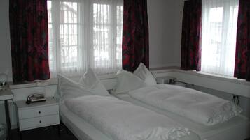 A double bed with white bedding in a room with two windows, white curtains, and a bedside table with a lamp and a telephone.