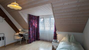 A bedroom with a sloped ceiling, a bed, a desk with a chair, a lamp, and a window with curtains.
