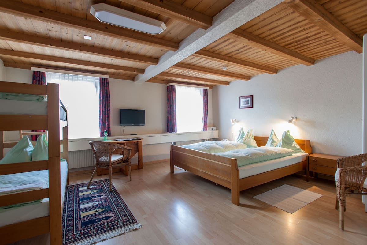 A room with a wooden ceiling, a bed, a desk with a chair, a TV, and a window with curtains.