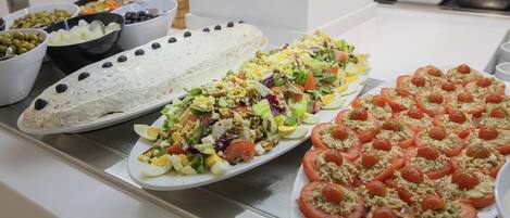 Un bufé con diversos platos, incluyendo una gran bandeja de tomates en rodajas cubiertos con una mezcla desmenuzada, una ensalada de hojas verdes y vegetales, y recipientes con aceitunas y otros productos.