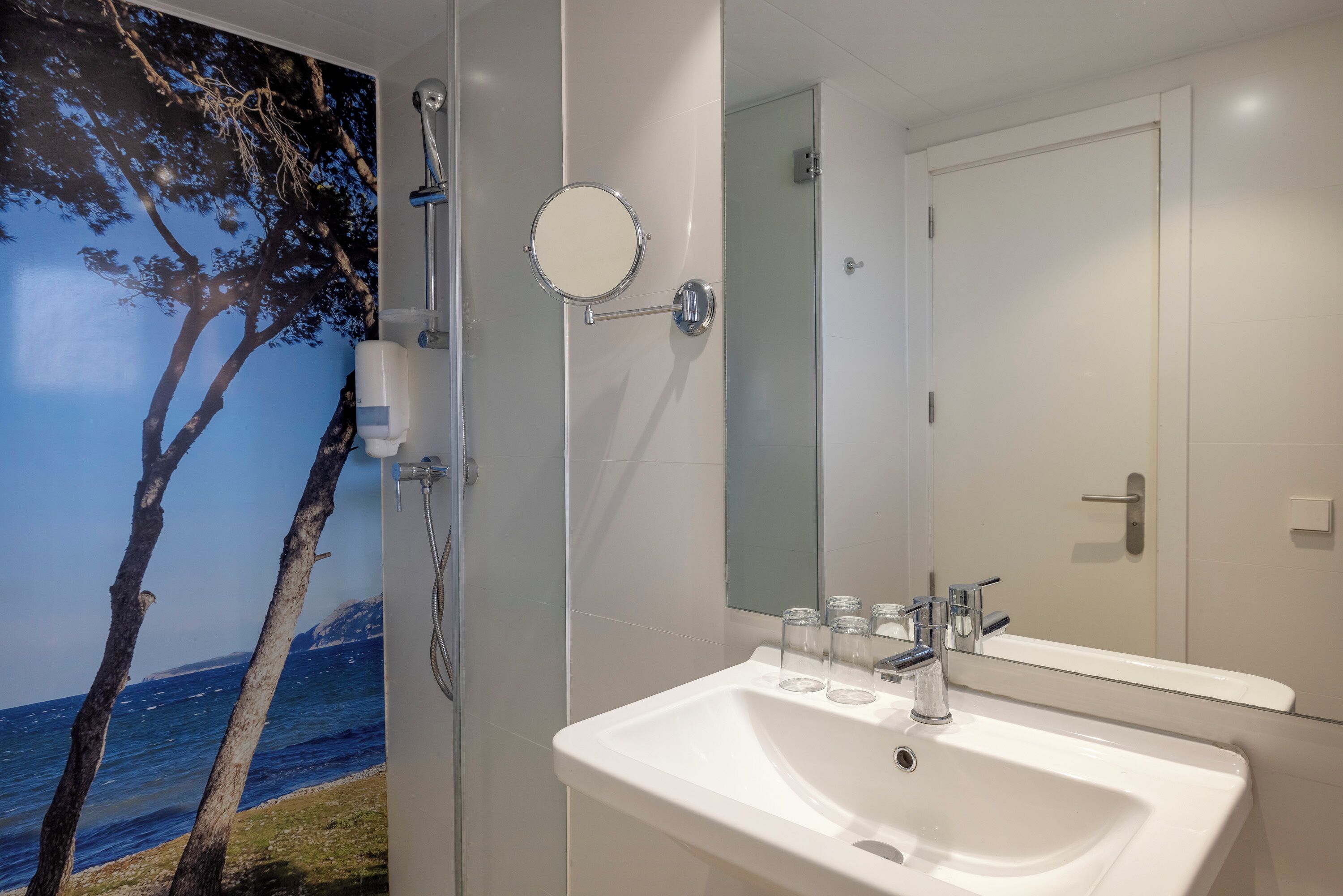 Un baño con lavamanos blanco, un espejo y una ducha. Hay un mural de un árbol en la pared.