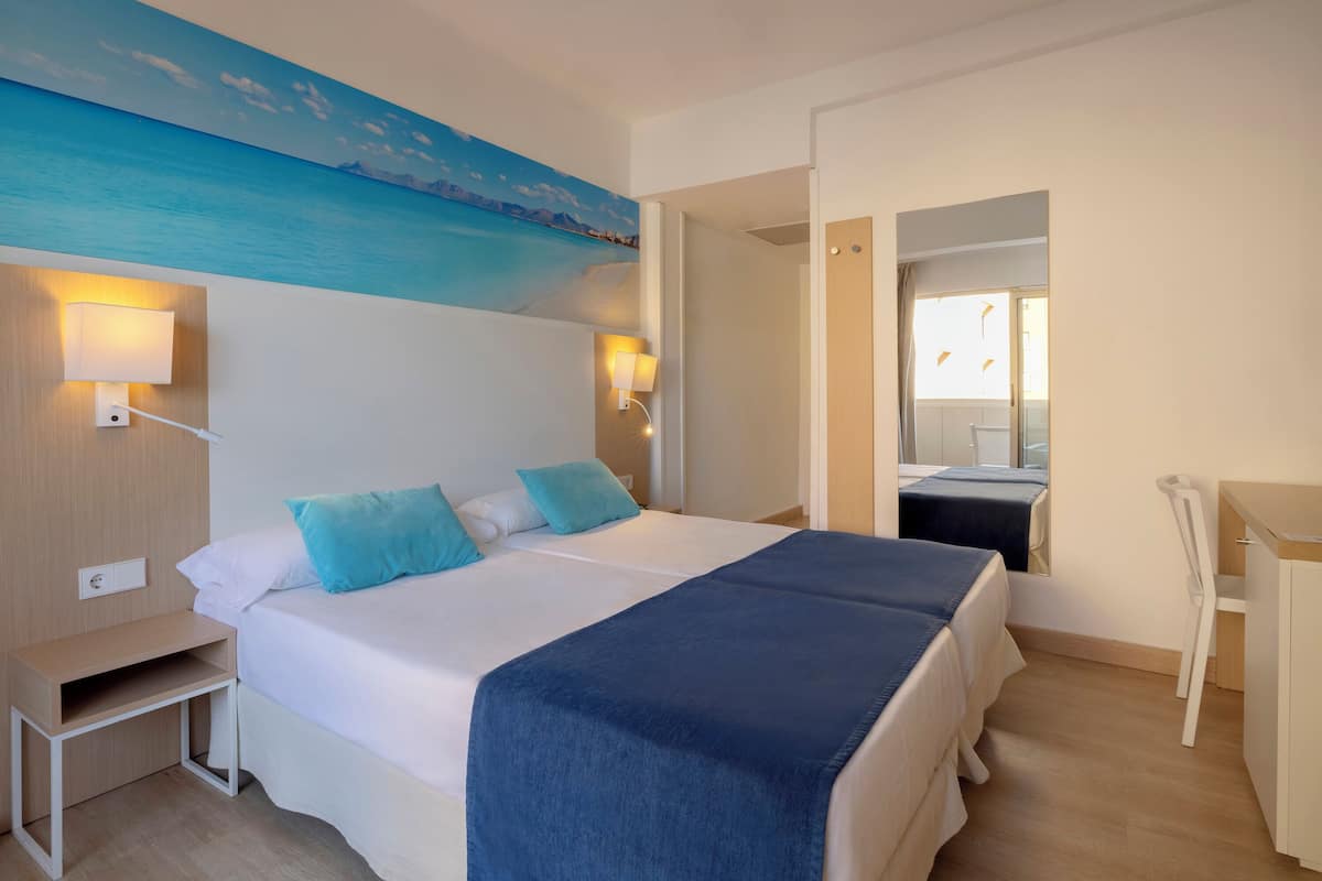 Ein Hotelzimmer mit Bett, Nachttischlampen, Schreibtisch und einem Bild von einer Strandlandschaft.