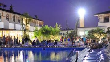 Zona de piscina nocturna con gente alrededor y edificios al fondo.