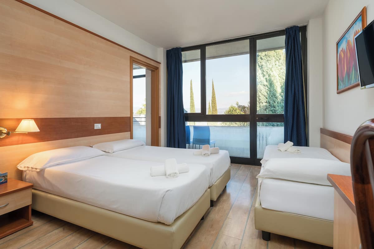 A hotel room with two beds, a TV, and a balcony with a view of trees.