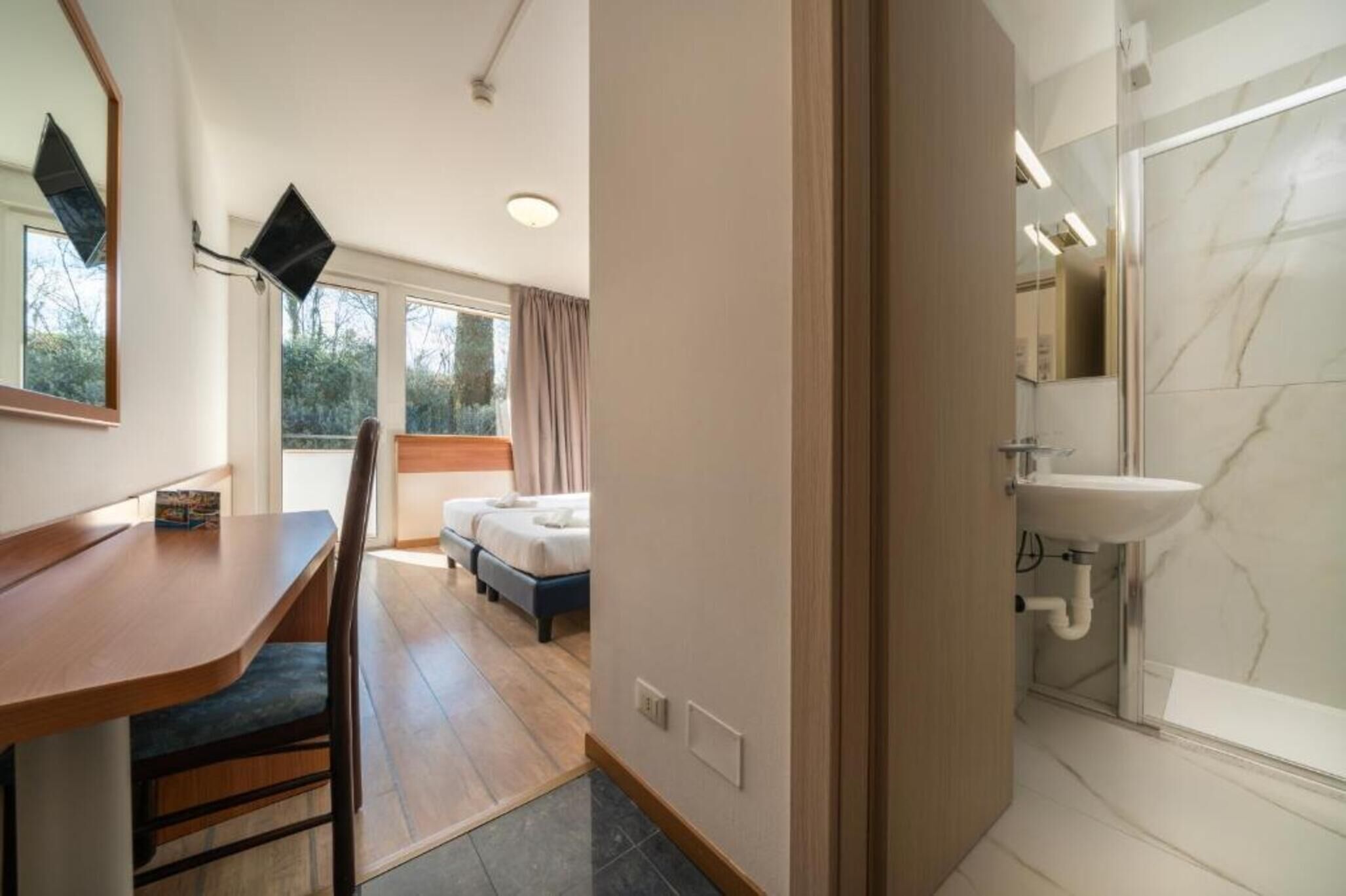 Habitación de hotel con un escritorio de madera, dos camas, un baño con lavamanos de mármol y ventanas grandes.
